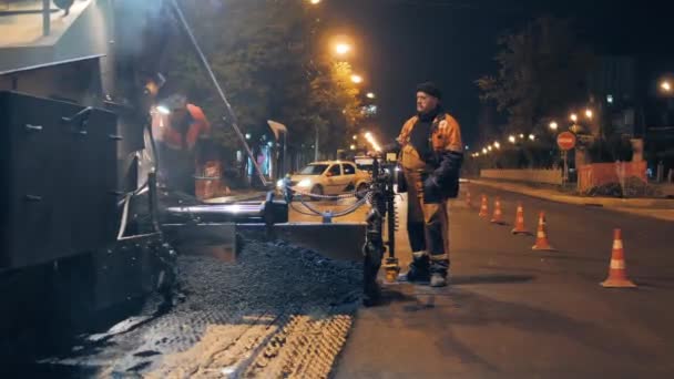 Novosibirská oblast, 7. září2019. Silniční dělník u ovládacího panelu asfaltových chodníků. Silniční oprava ve městě v noci. Auta jedou v pozadí. — Stock video