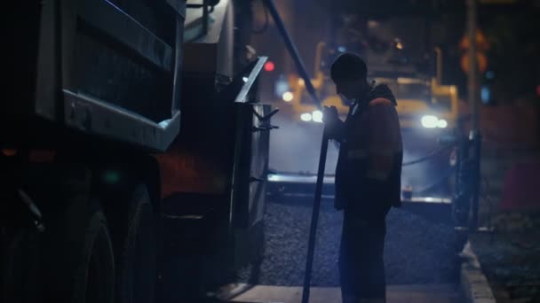 Novosibirsk região, 7 de setembro de 2019. Um trabalhador rodoviário supervisiona o carregamento de mistura de asfalto em um pavimentador de asfalto. Reparação de uma estrada de cidade à noite. — Vídeo de Stock