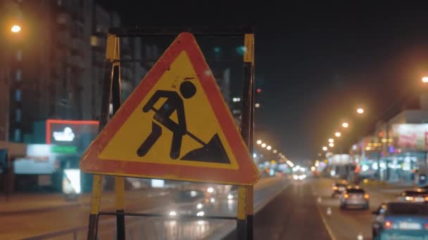 Primer plano de una señal de tráfico advirtiendo a los conductores sobre las obras viales. Señal de tráfico de Grunge. Contra el fondo de las luces de la ciudad nocturna y los coches que pasan. — Vídeos de Stock