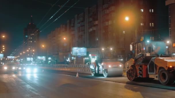 Obwód nowosybirski, 20 maja 2021 r. Trzy rolki drogowe układają asfalt na tle miasta w nocy. Samochody przejeżdżają. Światła nocnego miasta. Naprawa dróg. — Wideo stockowe