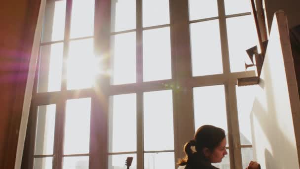Gebiet Nowosibirsk, 18. September 2020. Der Künstler malt auf einer Staffelei vor dem Hintergrund eines sonnigen Fensters. Sonnenstrahlen, Blendung. Kunstwerkstatt, Kunst, Kreativität. — Stockvideo