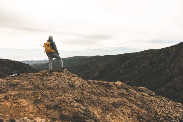 A man in hiking clothes with a yellow backpack stands on the top of a hill among the mountains around and in the background