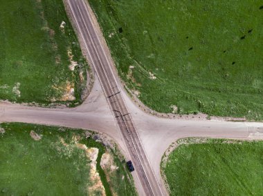 Üstten hava manzaralı yeşil çayırlar arasında iki kırsal karayolu kavşağı. Yalnız bir araba yolun kenarına park edilmiş.