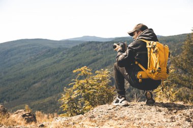 Sarı sırt çantalı bir adam dağlara tırmanıyor ve doğanın resimlerini çekiyor.