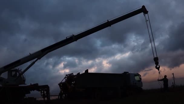 Silueta unei macarale de camion de lucru și un vehicul special pentru întreținerea puțurilor de petrol demontează echipamentul pe fundalul cerului de seară — Videoclip de stoc