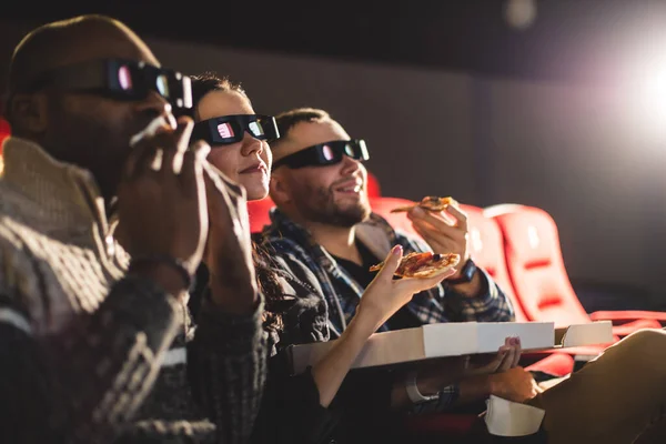 Friends watch a movie in the cinema, eat pizza and laugh. People sit in the armchairs of the cinema and look at the screen with 3D glass