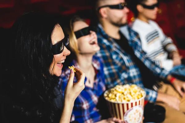 Friends watching a movie in the cinema with popcorn and laughing. People sit in the armchairs of the cinema and look at the screen with special glasses for 3D