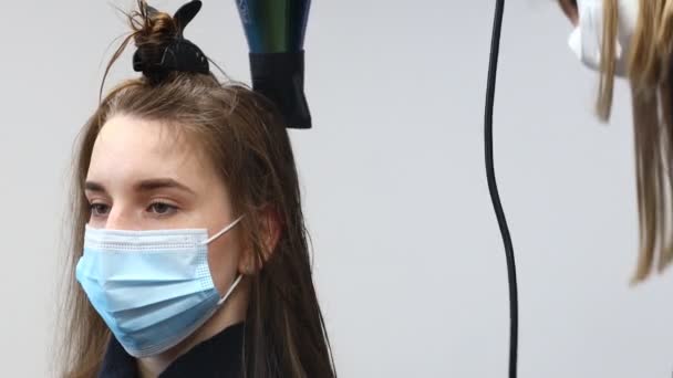 La peluquera maestra en una máscara médica seca el cabello de las niñas con un secador de pelo y peina después de lavarse en un salón de belleza. pandemia de Covid-19 y remedios — Vídeos de Stock