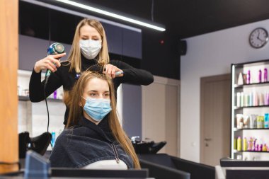 Tıbbi maskeli usta kadın kuaför bir güzellik salonunda yıkandıktan sonra saç kurutma makinesi ve tarak ile kızların saçlarını kurutuyor. Covid-19 salgını ve ilaçları