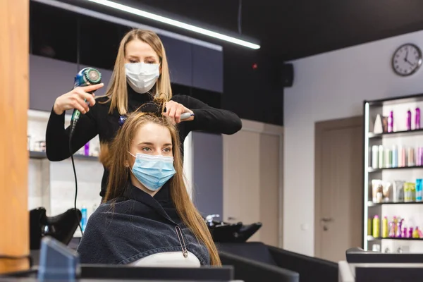 Master γυναίκα κομμώτρια σε ιατρική μάσκα στεγνώνει τα μαλλιά των κοριτσιών με στεγνωτήρα μαλλιών και χτένες μετά το πλύσιμο σε ένα σαλόνι ομορφιάς. Πανδημία και θεραπείες Covid-19 — Φωτογραφία Αρχείου