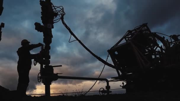 Um funcionário em macacão e um capacete está realizando reparos e manutenção de um poço de petróleo. Silhueta no fundo do céu da noite — Vídeo de Stock