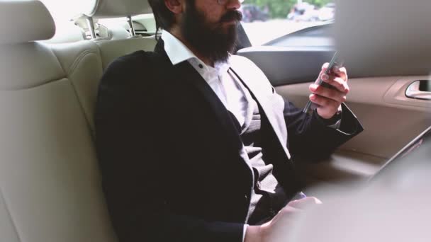 Індуський бізнесмен у костюмі і окулярах користується телефоном на задньому сидінні автомобіля. — стокове відео