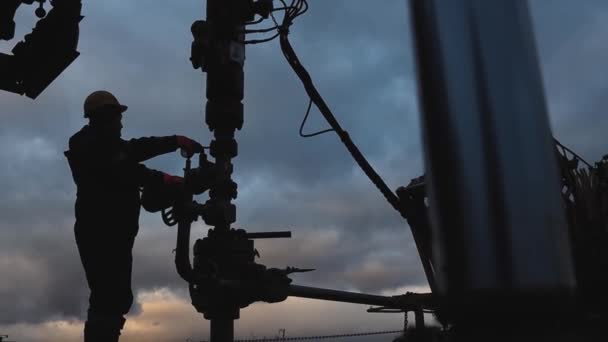 Een medewerker in overalls en een helm is bezig met reparaties en onderhoud van een oliebron. Silhouet op de achtergrond van de avondhemel — Stockvideo