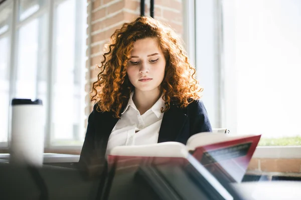 Retrato de una hermosa mujer de negocios joven con el pelo rizado rojo leyendo un libro en la oficina. Carrera exitosa y tiempo libre y aficiones — Foto de Stock