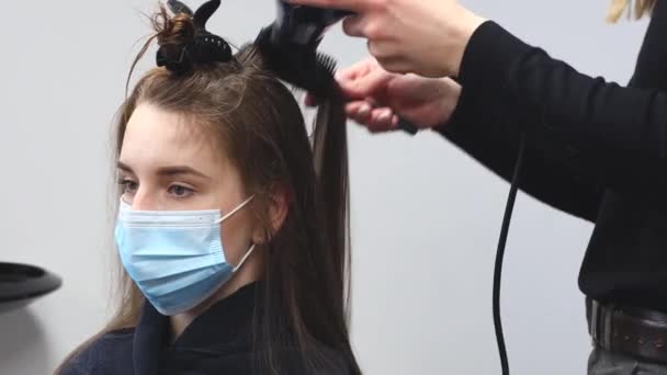 Il parrucchiere di donna di padrone in una maschera medica asciuga i capelli di ragazze con un asciugacapelli e pettini dopo essersi lavato in un salone di bellezza. Covid-19 pandemia e rimedi — Video Stock