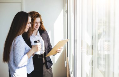 İki güzel genç iş kadını ofis penceresinin yanında iş konuşuyorlar ve tartışıyorlar..