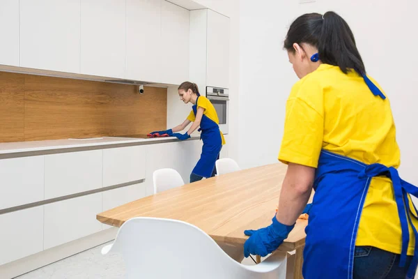 Servicio de limpieza profesional. Dos mujeres en uniforme de trabajo, en delantales, dividen la limpieza de la cocina de una casa privada, casa de campo. lavar el refrigerador, grifo, fregadero. Lavar el suelo. —  Fotos de Stock