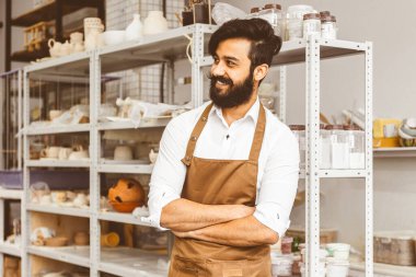 Atölyesinde sakalı ve bıyığı olan genç ve çekici bir işadamının portresi. Bitmiş ürünlerle rafların arka planında dururken çapraz pozlar vermek