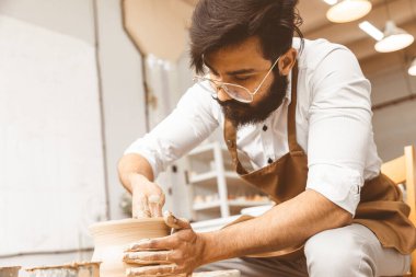 Genç bir erkek çömlekçi atölyesinde çömlekçi çarkında el işi yapıyor ve kilden bir ürün yapıyor.