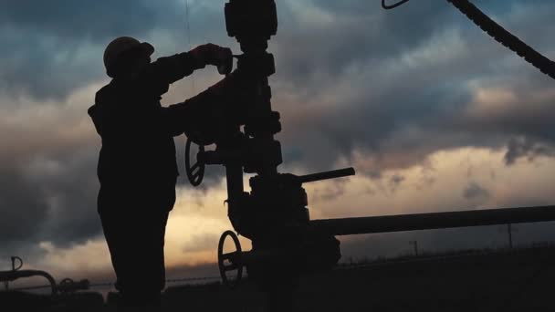 Un dipendente in tuta e un casco sta effettuando le riparazioni e la manutenzione di un pozzo petrolifero. Silhouette sullo sfondo del cielo serale — Video Stock