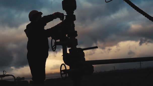 Um funcionário em macacão e um capacete está realizando reparos e manutenção de um poço de petróleo. Silhueta no fundo do céu da noite — Vídeo de Stock