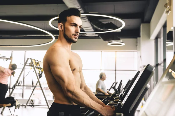 Jóképű fiatal sportos férfi testépítő meztelen felsőtesttel gyakorolja a futópadon az edzőteremben — Stock Fotó