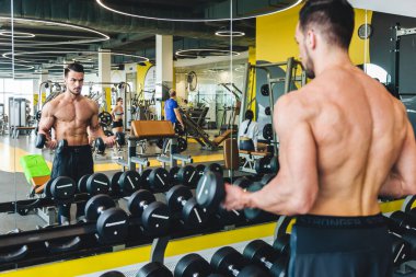 Spor salonunda, aynadan çıplak bir şekilde dambıl egzersizi yapan sporcu.