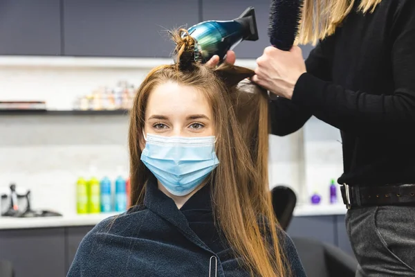 Master kobieta fryzjer w masce medycznej suszy włosy dziewcząt z suszarką do włosów i grzebienie po umyciu w salonie piękności. Pandemia i środki zaradcze przeciwko chorobie Covida-19 — Zdjęcie stockowe