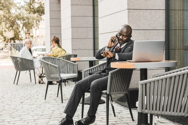 Porträtt av en svart afroamerikansk affärsman i kostym som sitter på ett kafé utomhus och pratar i telefon. — Stockfoto
