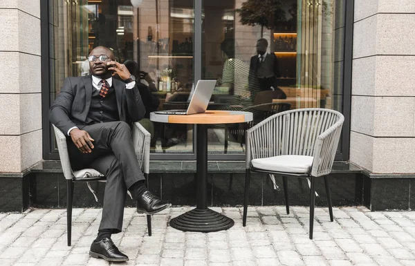 Retrato de un hombre de negocios afroamericano en traje sentado en un café de la ciudad al aire libre y hablando por teléfono. — Foto de Stock