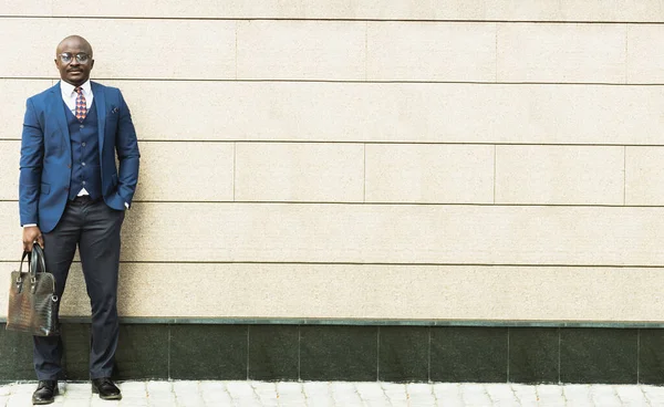 Retrato de cuerpo entero de hombre de negocios afroamericano negro en traje y gafas al aire libre con maletín en la mano contra la fachada del edificio fondo de la pared — Foto de Stock