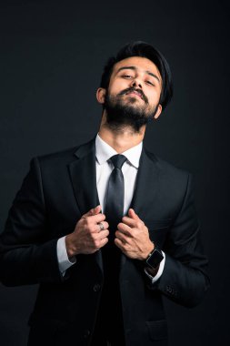 Siyah arka planda üç parçalı klasik bir iş elbisesi içinde iki eliyle ceketin yakasını tutan yakışıklı ve başarılı bir Hintlinin portresi.