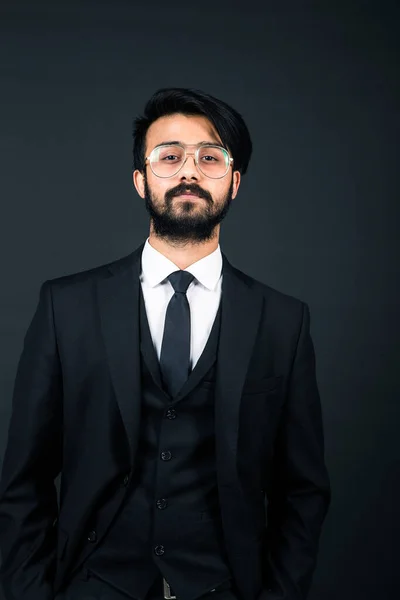 Portret van een jonge knappe succesvolle Indiaan in een klassiek zwart driedelig zakenpak op een donkere achtergrond, houd de bril naar beneden — Stockfoto