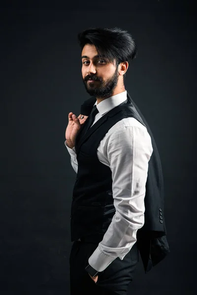 Retrato de um jovem indiano bonito e bem sucedido em um terno de negócios preto clássico de três peças em um fundo escuro, segurando seu casaco sobre seu ombro — Fotografia de Stock
