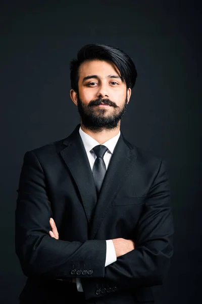 Portret van een jonge knappe succesvolle Indiaan in een klassiek zwart driedelig zakenpak op een donkere achtergrond. armen gekruist — Stockfoto
