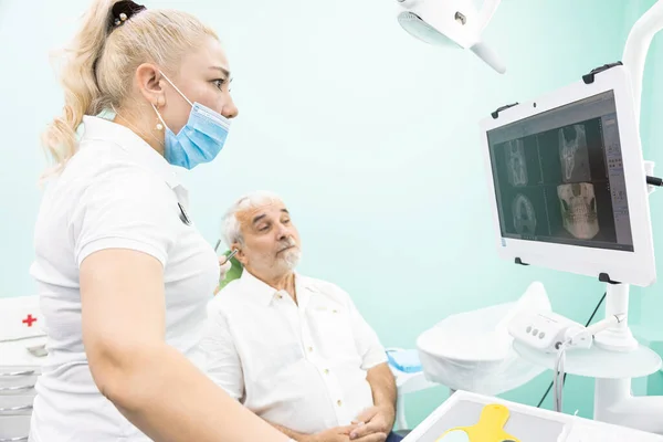 Odontologia e saúde. Serviços odontológicos profissionais e equipamentos modernos sem dor. O médico consulta um paciente idoso, olha para as fotos dos dentes na tela do monitor — Fotografia de Stock