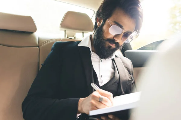 Un indio elegante y exitoso con gafas y barba trabaja y planea su horario en el asiento trasero del coche. Viajes de negocios seguros y cómodos — Foto de Stock