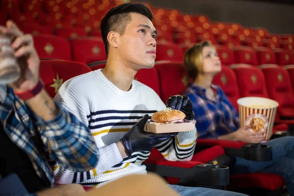 Amigos viendo una película en el cine y comiendo hamburguesas. La gente se sienta en los sillones del cine y mira la pantalla — Foto de Stock