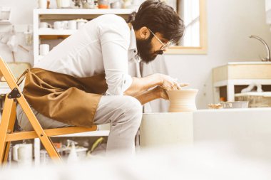 Genç bir erkek çömlekçi atölyesinde çömlekçi çarkında el işi yapıyor ve kilden bir ürün yapıyor.