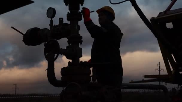 Een medewerker in overalls en een helm is bezig met reparaties en onderhoud van een oliebron. Silhouet op de achtergrond van de avondhemel — Stockvideo