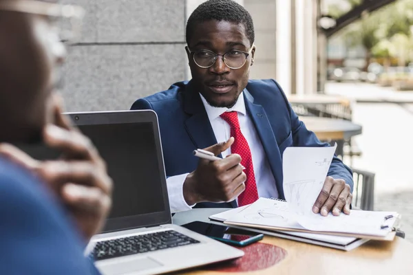 Två mörkhyade afroamerikanska affärsmän i kostym och glasögon med portföljer sitter vid ett bord på ett uteservering. — Stockfoto