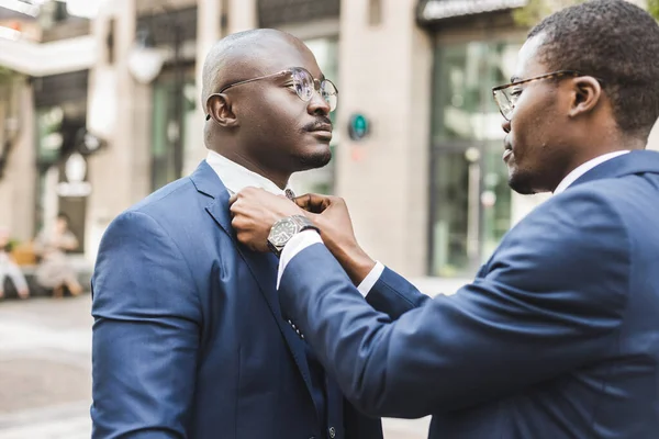 Two black African American businessman friends before an important business meeting. One helps the other to put on a tie and straightens the suit