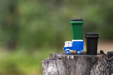 Oyuncak çöp kamyonu ve çöp tenekeleri bulanık orman arka planına karşı ağaç keneviri üzerinde duruyor. Seçici odak, uzayı kopyala. Ormanları temiz tutma kavramı.