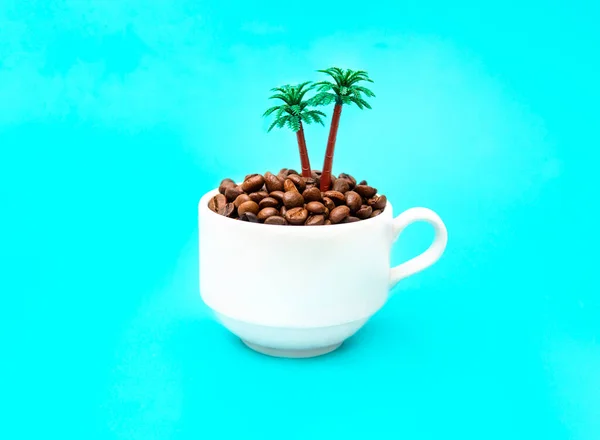 白咖啡杯 满是烤咖啡豆和玩具棕榈树 背景浅蓝色 具有创意的偏远岛屿概念 — 图库照片
