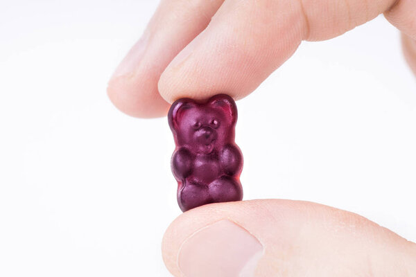 Crop view of male fingers holding a purple gummy bear against white background.