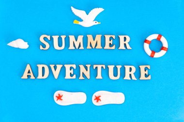 Yaratıcı yaz tatili kompozisyonu: Oyuncak martı, parmak arası terlik, can simidi ve deniz kabuğuyla birlikte mavi arkaplandaki ahşap karakterlerden oluşan bir harf..