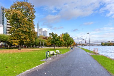Frankfurt, Germany - October 21, 2025: Lush autumn parkland beside Frankfurt's Main River, providing a tranquil promenade with benches and a modern city skyline vista.