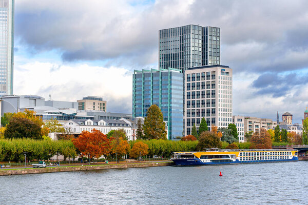 Франкфурт, Германия - 21 октября 2025 года: вид на набережную Франкфурта-на-Майне с современными небоскребами, жилыми зданиями и яркими осенними деревьями, с круизным кораблем на воде.