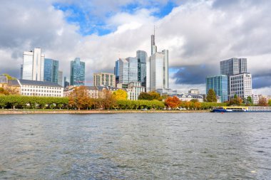 Frankfurt, Almanya - 21 Ekim 2025: Frankfurt 'un modern finans bölgesi, canlı sonbahar ağaçları ve geçen bir nehir teknesi tarafından çerçevelenen Main River' ın üzerinde görkemli bir şekilde yükseliyor.