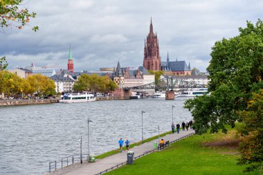 Frankfurt, Almanya - 21 Ekim 2025: Kentsel nehir manzarası, Frankfurt şehrinin tarihi mimari unsurlarıyla yan yana eğlence amaçlı koşu ve yürüyüş yapan bireyleri tasvir ediyor.